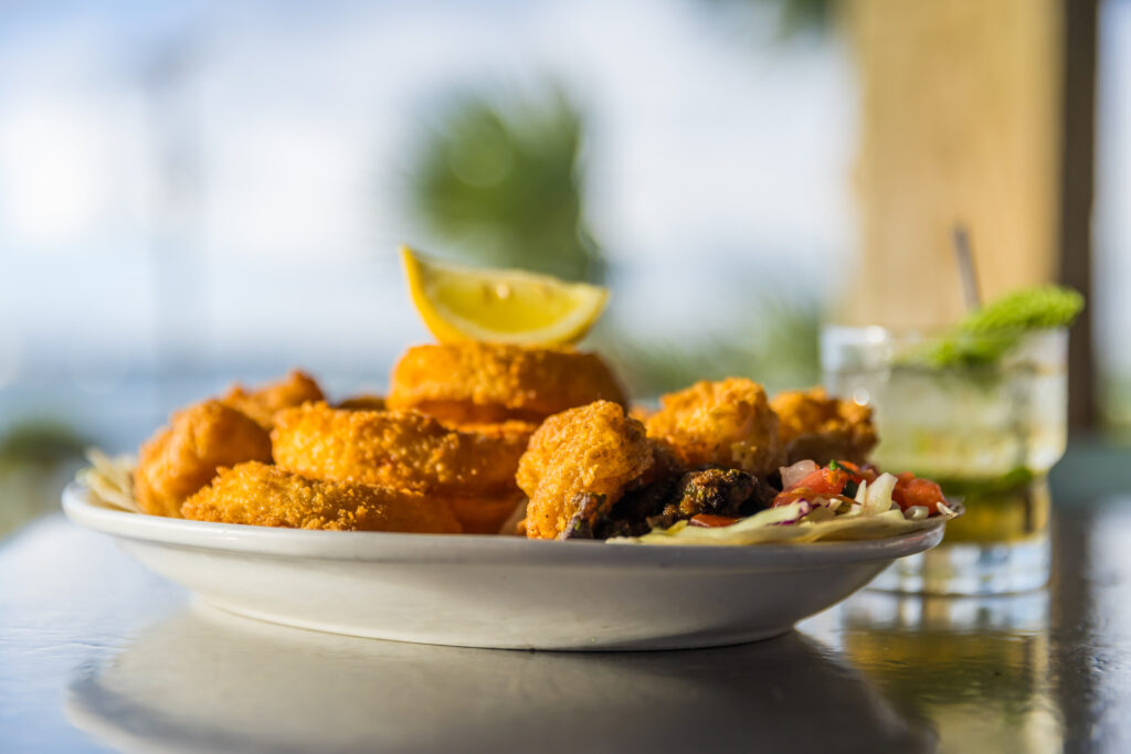 A plate of golden fried shrimp and onion rings garnished with a lemon wedge, served with a side of coleslaw. A cocktail with mint is in the background on a reflective table.