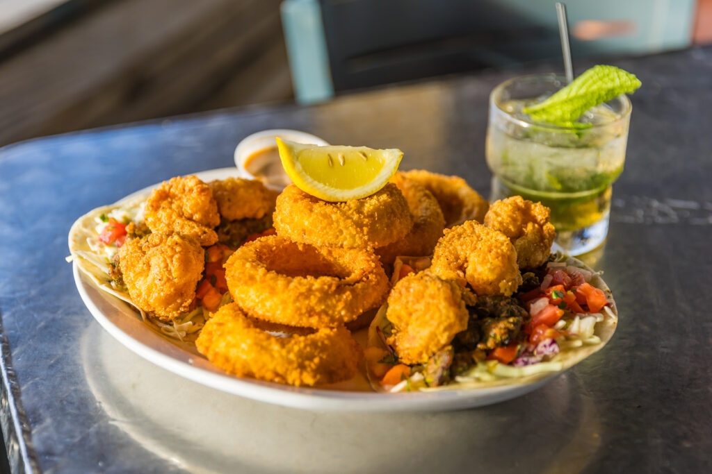 A plate of golden fried shrimp and onion rings garnished with a lemon wedge, served on a bed of shredded vegetables with a dipping sauce, next to a glass of cocktail with mint on a reflective table.