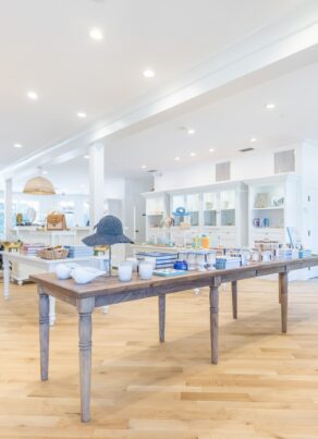 A bright, modern retail store with wooden floors and white shelves displaying various products. In the center, a wooden table holds ceramic items and decor. The space is well-lit with ceiling lights.
