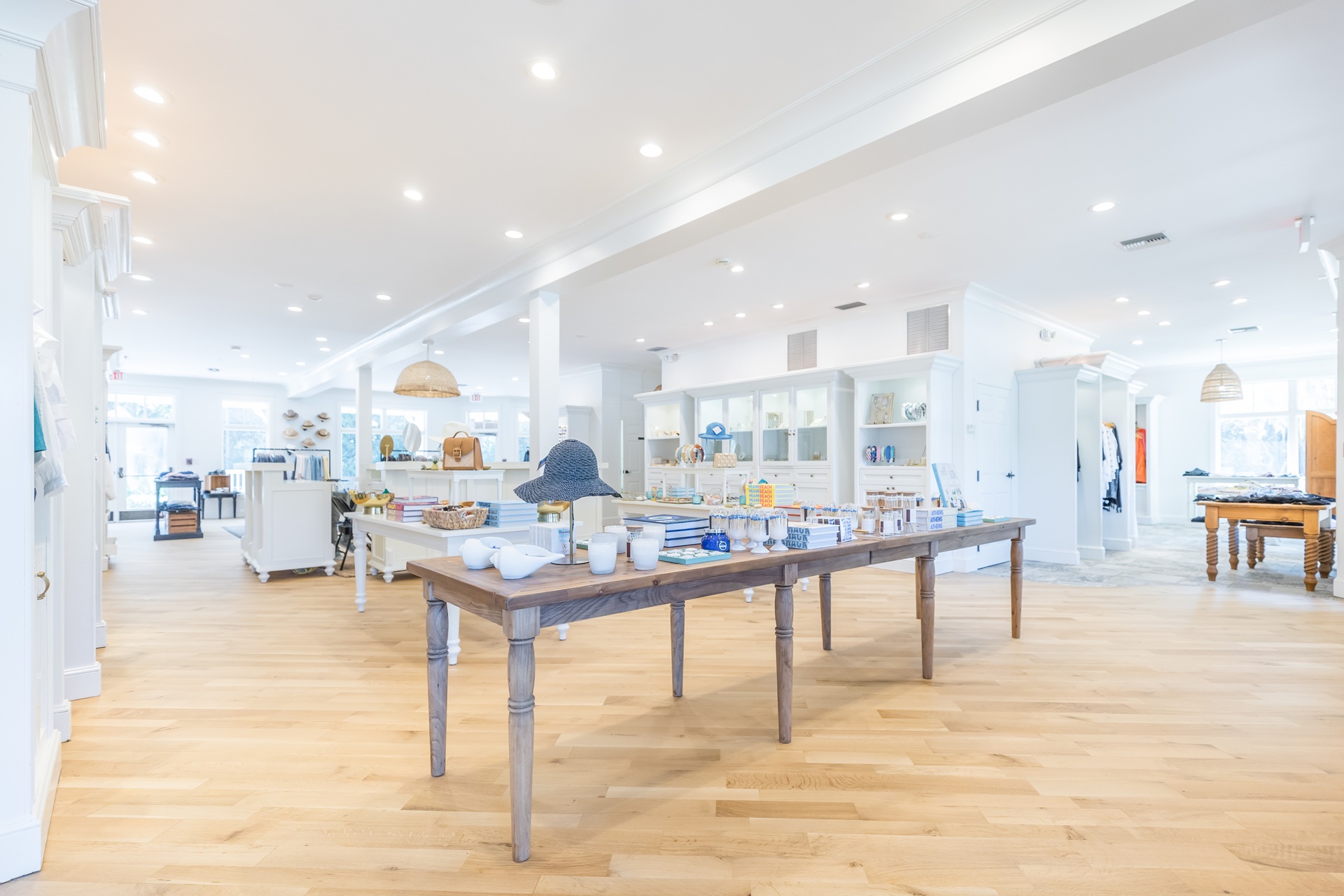 A bright, modern retail store with wooden floors and white shelves displaying various products. In the center, a wooden table holds ceramic items and decor. The space is well-lit with ceiling lights.