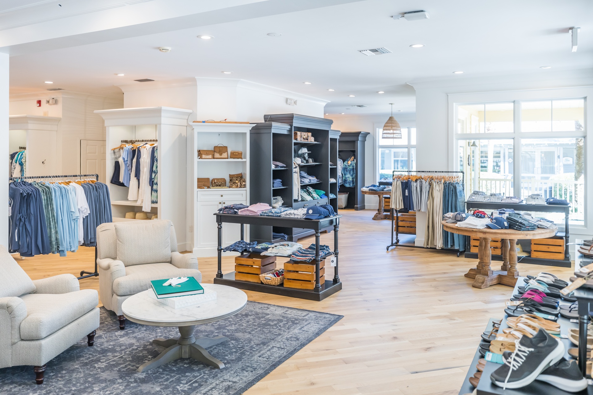 A bright, modern clothing store with wooden floors, racks of mens clothes, shelves with folded shirts, casual shoes on display, and two cushioned chairs with a round table in the foreground.
