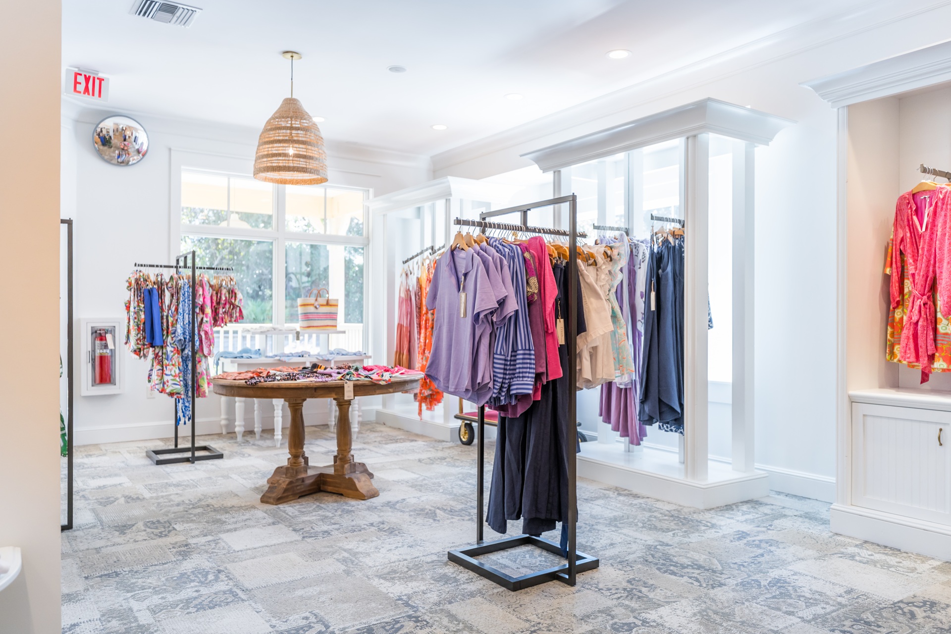 A bright boutique with hanging racks of colorful womens clothing, a round wooden display table in the center, large windows, and white walls with natural light filling the space.