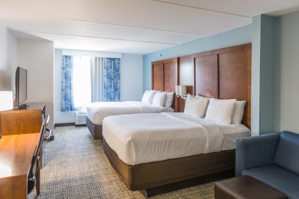 A hotel room with two queen beds, white bedding, wooden headboards, a desk with a chair, a TV, blue curtains over the window, and a blue sofa in the corner. The room has neutral walls and carpeted flooring.