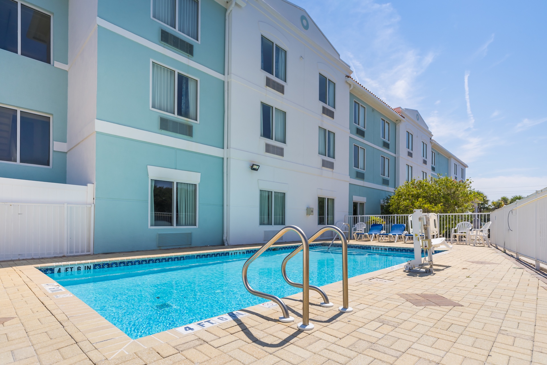 A small outdoor swimming pool with metal handrails sits beside a three-story hotel building. Several lounge chairs and a pool lift are on the surrounding paved area under a sunny blue sky.