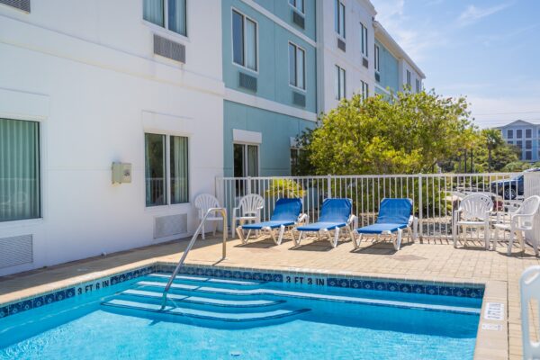 Outdoor pool with blue water, steps leading in, and a metal handrail. Four blue lounge chairs and three white plastic chairs are arranged on the pool deck, next to a white fence and a building with pale blue and white walls.
