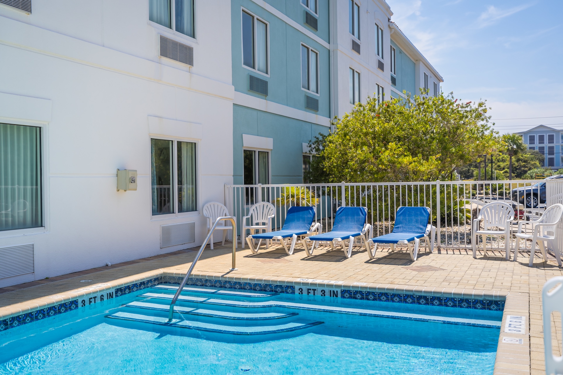 Outdoor pool with blue water, steps leading in, and a metal handrail. Four blue lounge chairs and three white plastic chairs are arranged on the pool deck, next to a white fence and a building with pale blue and white walls.