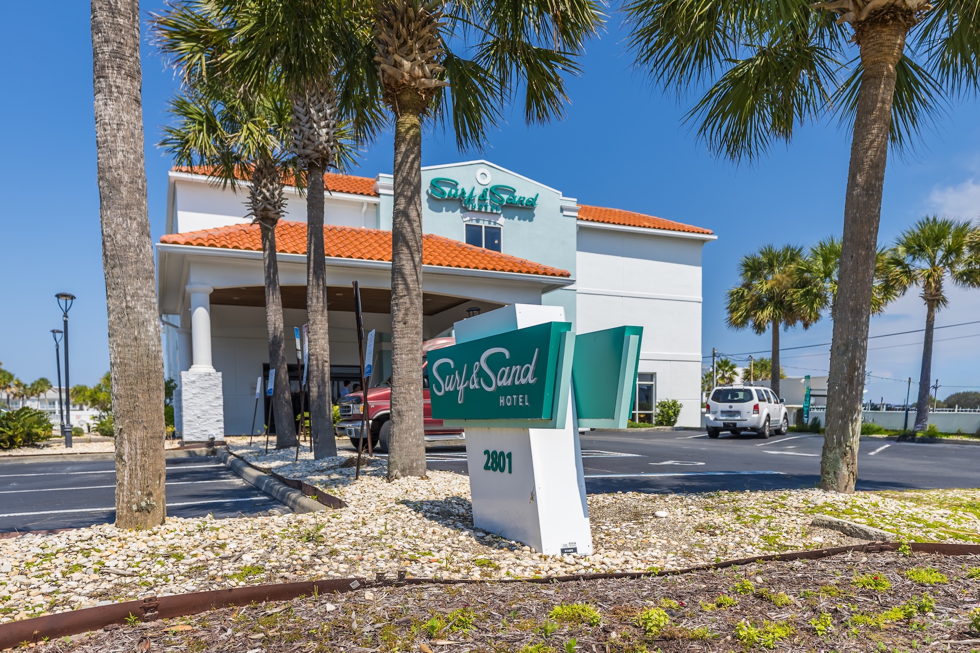A hotel with a red-tiled roof and palm trees in front, featuring a green and white sign that reads Sugar Sands Hotel 2801. A white SUV is parked in the lot under a clear blue sky.