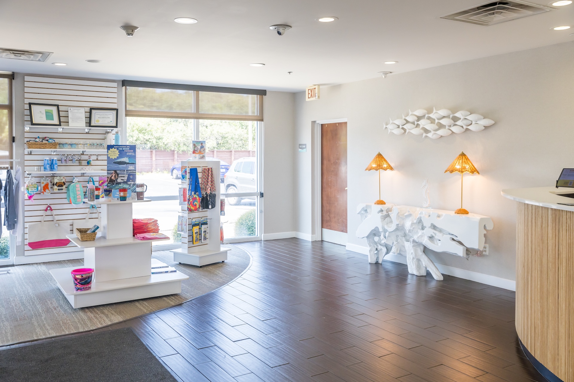 A bright, modern lobby with dark wood floors, a curved front desk, retail shelves displaying pet products, a white table with two orange lamps, and large windows letting in natural light.
