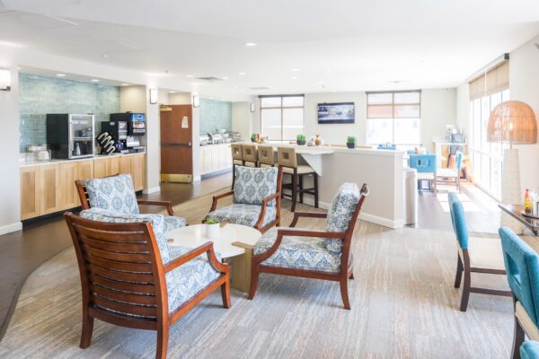 Bright, modern hotel breakfast area with wood and upholstered chairs around tables, a breakfast counter with food and drink stations, and large windows letting in natural light.