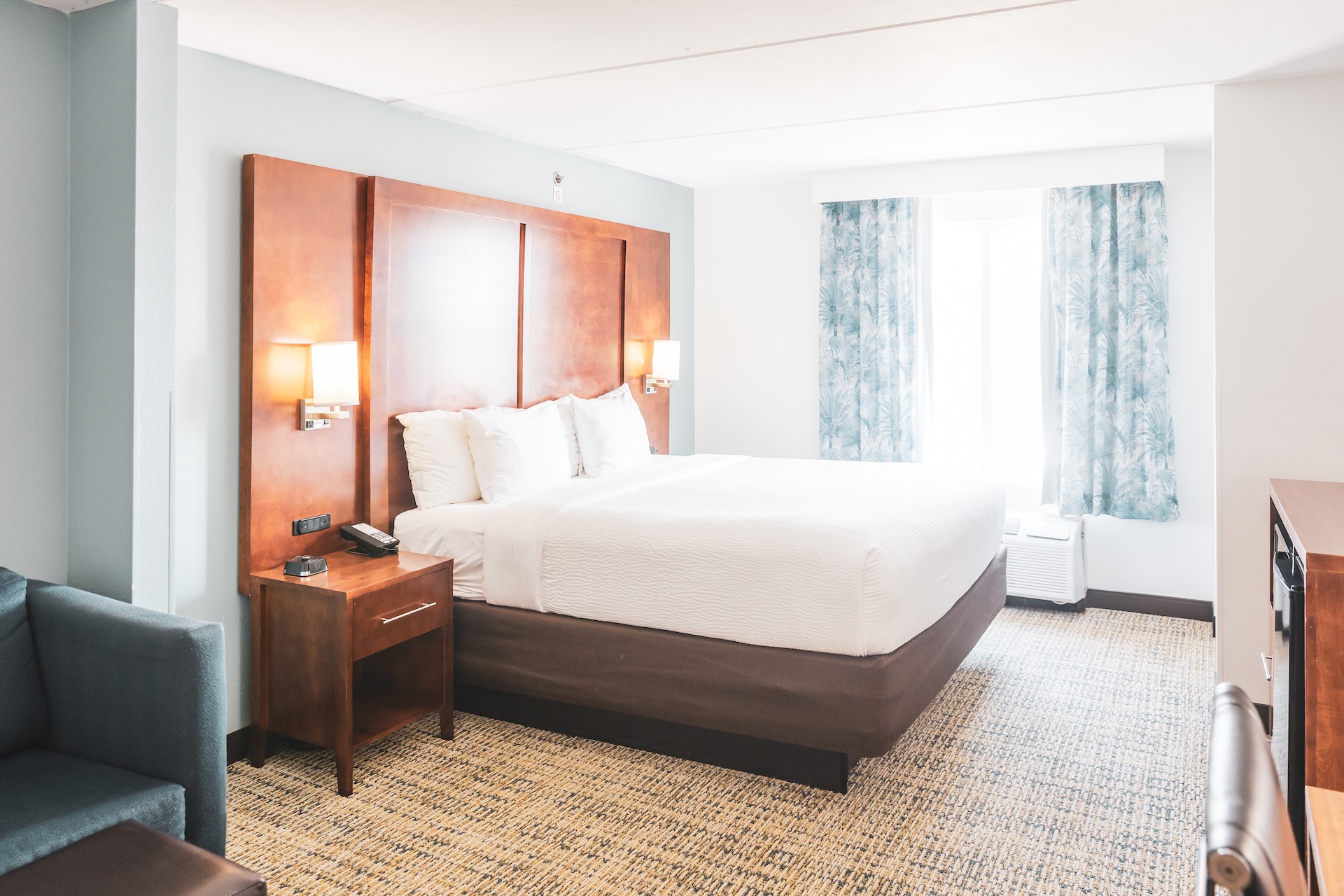 A hotel room with a neatly made king-size bed, wooden headboard, bedside tables with lamps, a sofa, patterned carpet, and a window with blue curtains letting in natural light.