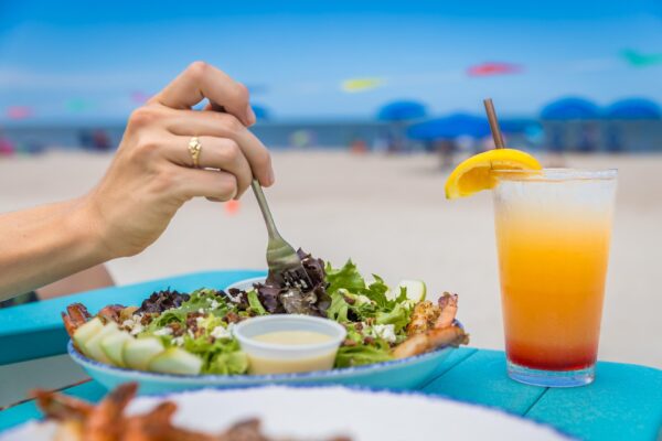 A hand holds a fork over a fresh salad in a bowl beside a tropical drink with an orange slice, set on a blue table at a beach with sand and blue umbrellas in the background.