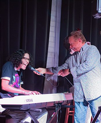 A person plays a Yamaha keyboard and sings into a microphone held by another smiling person on stage, with dark curtains in the background.
