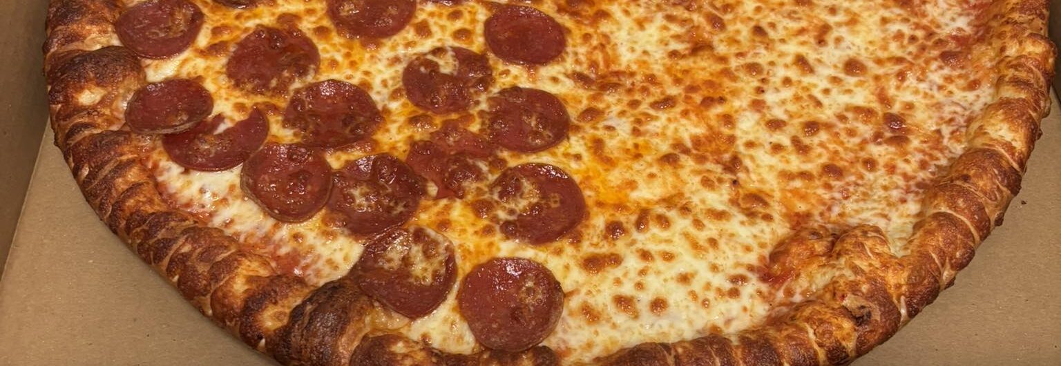 A large pizza in an open cardboard box sits on a metal counter. Half the pizza is topped with pepperoni slices, while the other half is plain cheese.