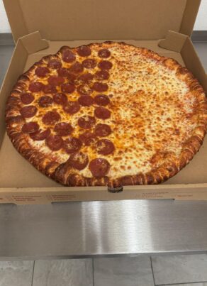A large pizza in an open cardboard box sits on a metal counter. Half the pizza is topped with pepperoni slices, while the other half is plain cheese.