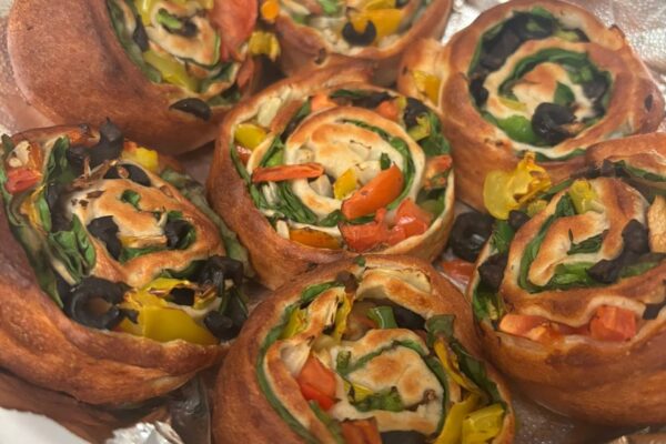 Seven baked pinwheels filled with spinach, sliced bell peppers, tomatoes, and black olives, placed on foil. The pastry is golden brown and the vegetables are colorful and fresh.