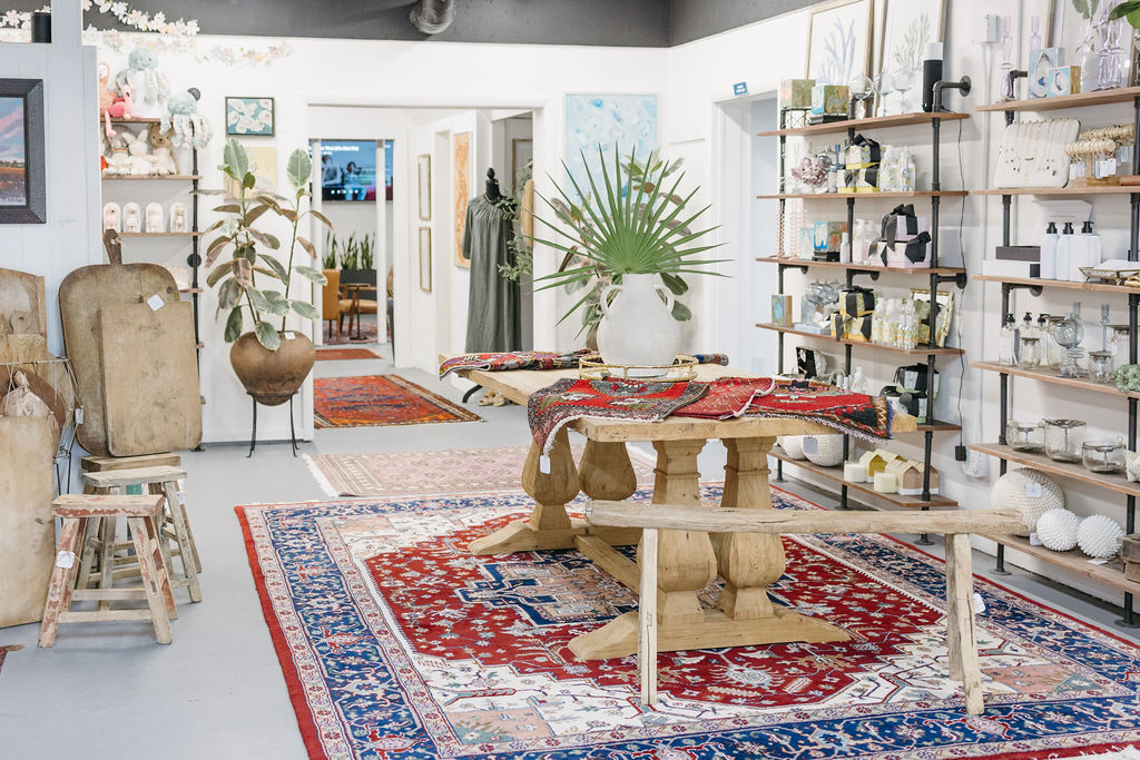 A bright boutique features wooden furniture, colorful patterned rugs, shelves with decorative items, potted plants, and framed artwork displayed on white walls. A large green plant in a vase sits on a central table.