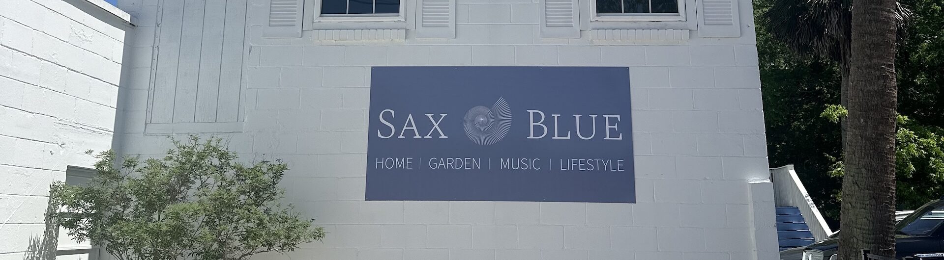 A white building with two striped blue-and-white awnings over windows. A sign reads Sax Blue: Home, Garden, Music, Lifestyle. A potted plant sits by the wall on a sunny day.
