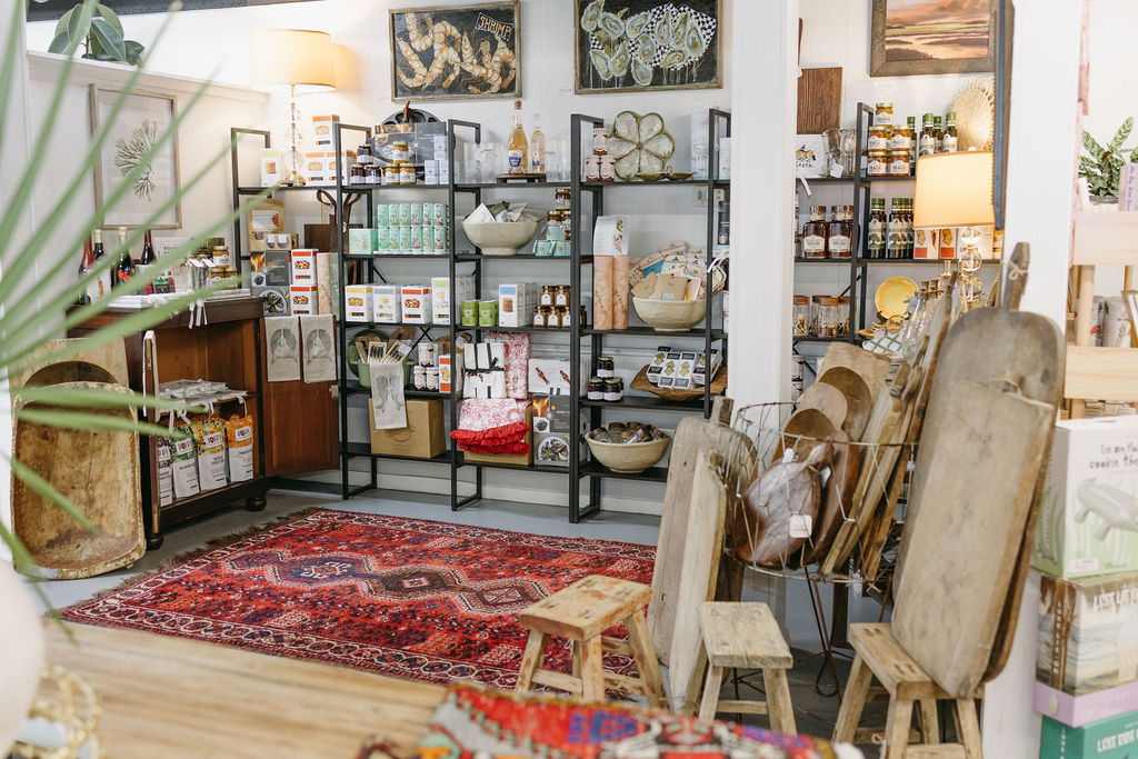 A cozy shop displays shelves of artisanal goods, kitchen items, and decor. Wooden stools and boards sit near a vibrant red patterned rug, with plants and framed art adding a warm, inviting touch to the scene.