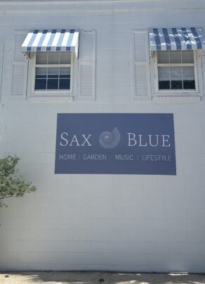 A white building with two striped blue-and-white awnings over windows. A sign reads Sax Blue: Home, Garden, Music, Lifestyle. A potted plant sits by the wall on a sunny day.