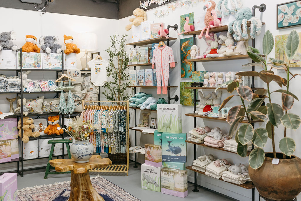 A boutique baby store displays stuffed animals, soft blankets, baby clothes on hangers, books, and various gifts on shelves. Plants and decorative items accent the cozy, well-lit space.
