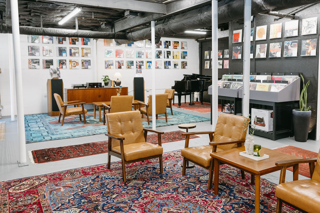 A stylish, retro-themed room with leather chairs, wooden tables, colorful rugs, a piano, and walls lined with vinyl records and album covers, creating a cozy, vintage lounge atmosphere.