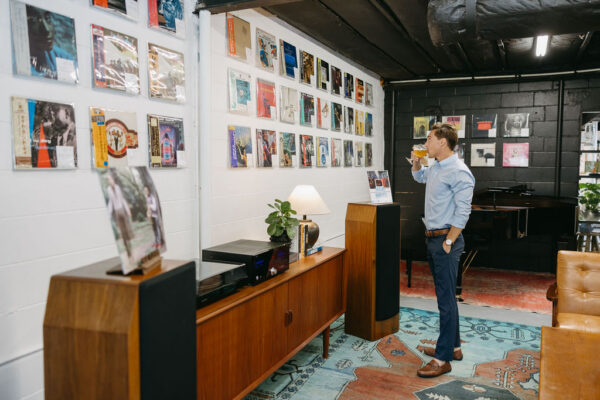 A man stands in a stylish room with a drink, looking at a wall display of vinyl records. The room has mid-century modern furniture, a record player, and decorative plants.