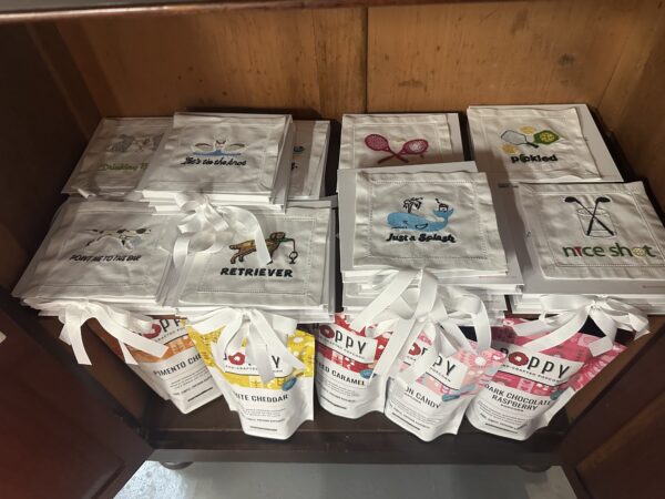 A wooden shelf displays stacks of white cloth napkins with playful embroidered designs and text, alongside packets of flavored popcorn from the brand Poppy on the lower shelf.