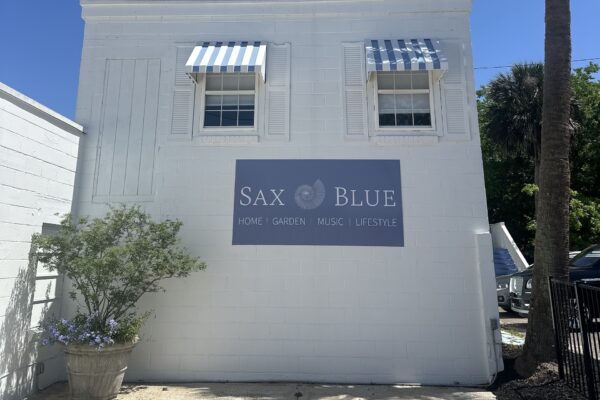 A white building with two striped blue-and-white awnings over windows. A sign reads Sax Blue: Home, Garden, Music, Lifestyle. A potted plant sits by the wall on a sunny day.