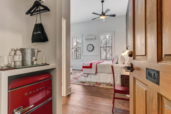 A cozy bedroom with a red retro mini fridge, a bed with white linens and red accents, a wooden desk and chair, a ceiling fan, and large windows letting in natural light. A door marked No. 8 is partially open.