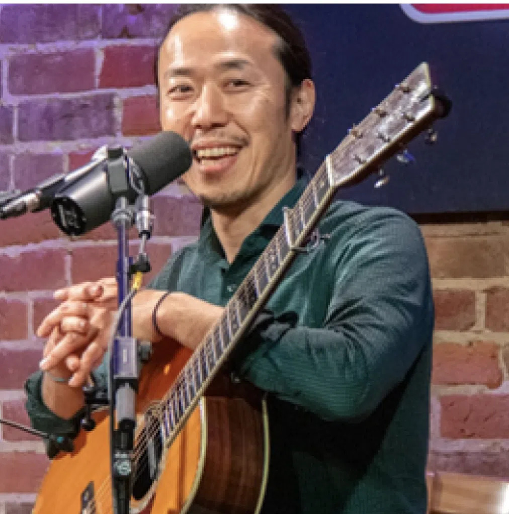A smiling man holding an acoustic guitar stands in front of a microphone.