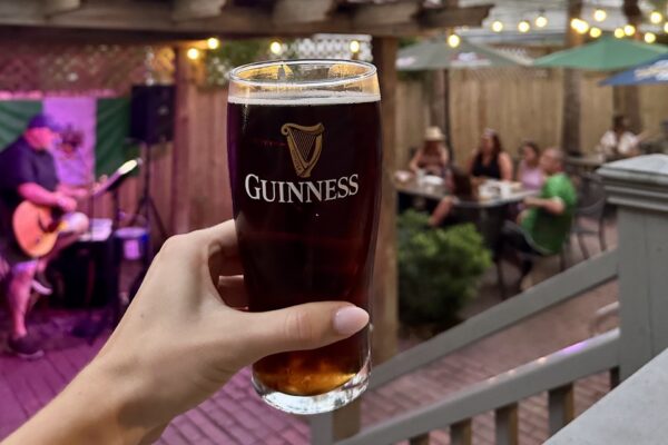 A hand holds up a pint of dark Guinness beer in an outdoor patio setting, with people sitting at tables and a musician playing guitar in the background under string lights.
