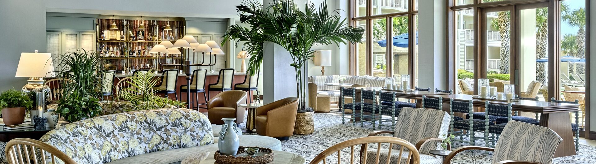 Bright, airy lounge with wooden beams, patterned flooring, a mix of cozy chairs, sofas, plants, and tables. Large windows overlook outdoor seating and greenery. Stylish, modern, and inviting space.