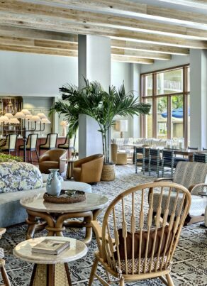 Bright, airy lounge with wooden beams, patterned flooring, a mix of cozy chairs, sofas, plants, and tables. Large windows overlook outdoor seating and greenery. Stylish, modern, and inviting space.
