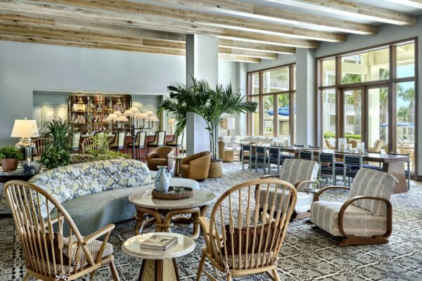 Bright, airy lounge with wooden beams, patterned flooring, a mix of cozy chairs, sofas, plants, and tables. Large windows overlook outdoor seating and greenery. Stylish, modern, and inviting space.