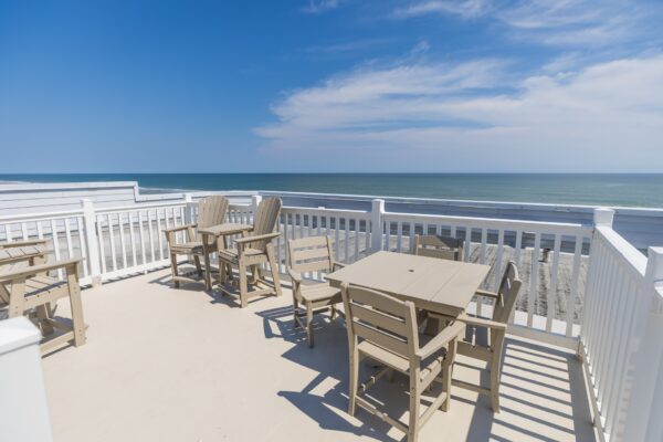 Outdoor deck with light-colored wooden tables and chairs overlooking a sandy beach and the ocean, under a sunny blue sky with scattered clouds. White railings surround the deck area.