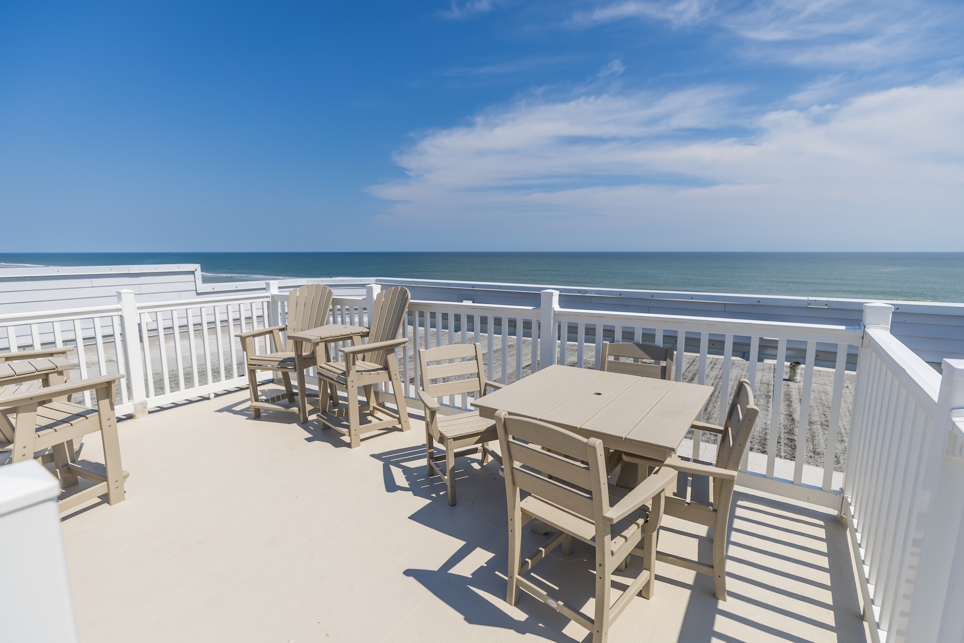 Outdoor deck with light-colored wooden tables and chairs overlooking a sandy beach and the ocean, under a sunny blue sky with scattered clouds. White railings surround the deck area.