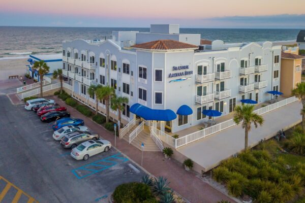 A beachfront hotel with blue awnings, balconies, and palm trees in front, overlooking a parking lot filled with cars, with the ocean and sandy beach visible in the background at sunset.
