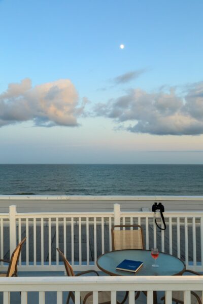 A round glass table with a book and a wine glass sits on a deck overlooking the ocean. Empty chairs surround the table, and a camera hangs on the white railing. The sky is blue with clouds and the moon is visible.