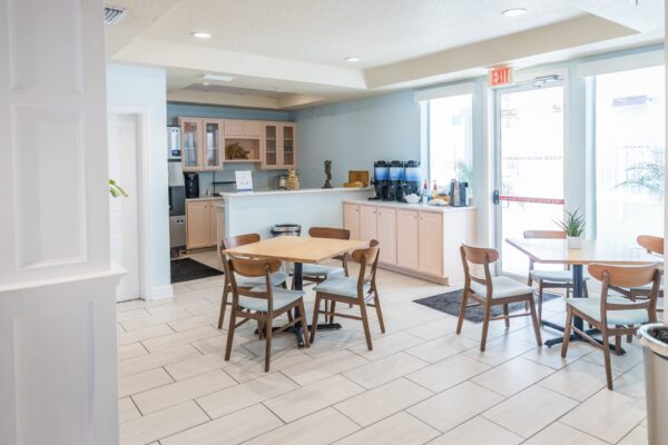 A small, bright cafe area with wooden tables and chairs, a coffee and beverage station, light-colored tile flooring, and large windows letting in natural light.