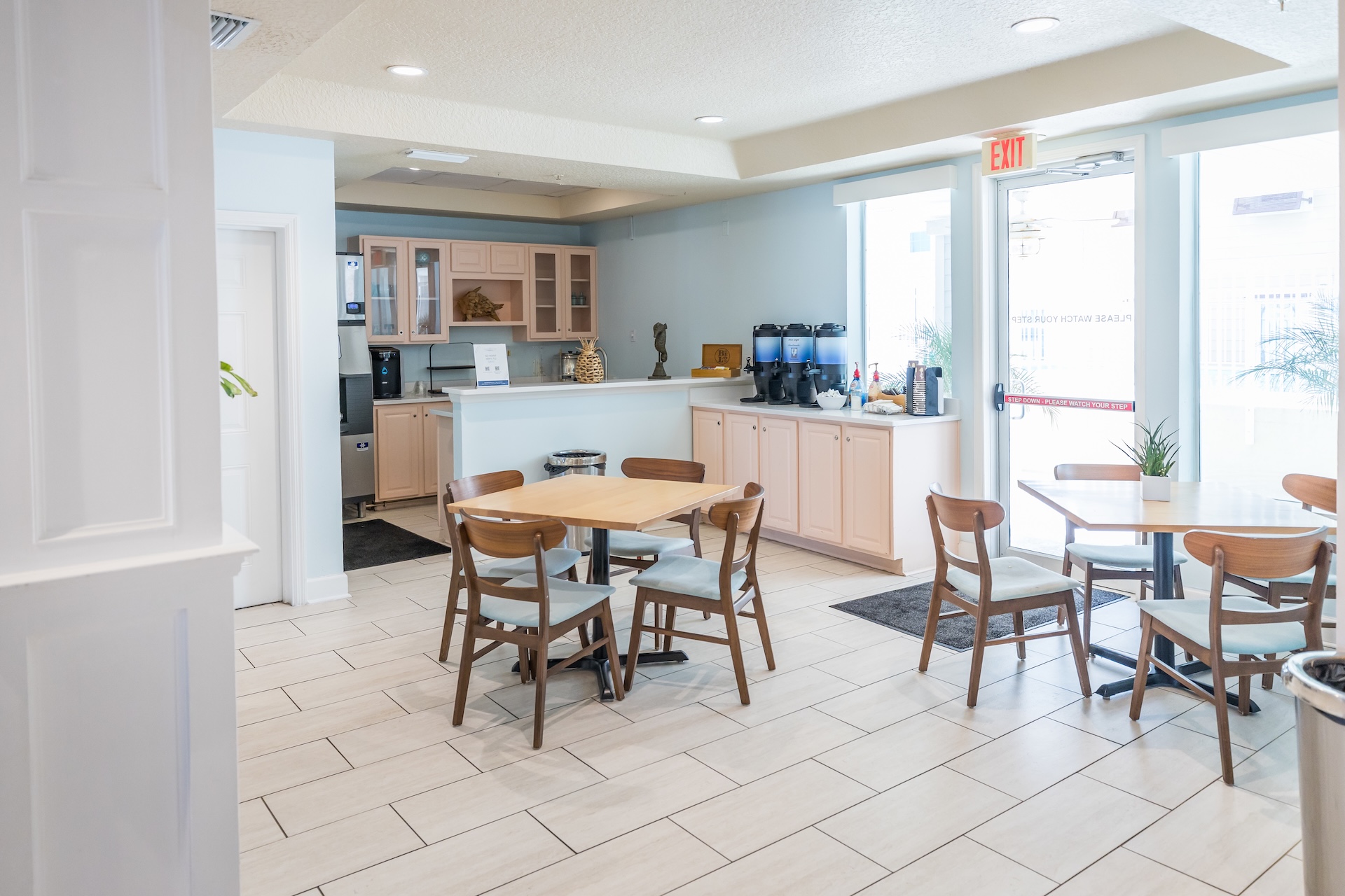 A small, bright cafe area with wooden tables and chairs, a coffee and beverage station, light-colored tile flooring, and large windows letting in natural light.