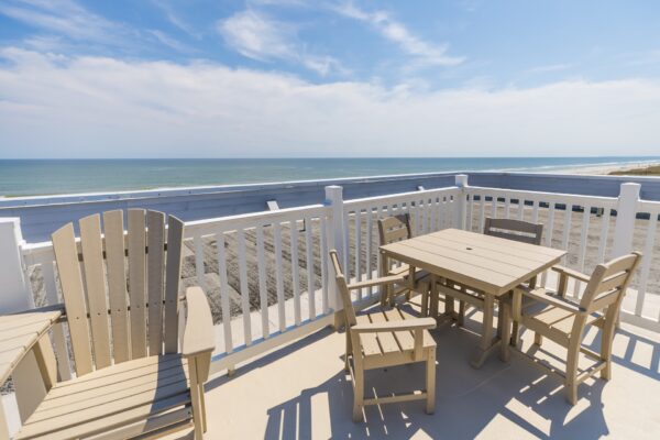A rooftop patio with wooden chairs and a table overlooks a sandy beach and calm ocean under a partly cloudy sky. White railings enclose the patio, offering a clear, scenic coastal view.
