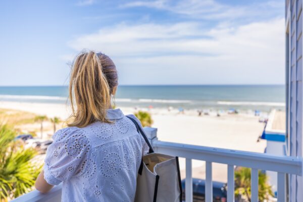 A woman with blonde hair in a ponytail, wearing a white eyelet shirt and carrying a tote bag, stands on a balcony overlooking a sandy beach and the ocean under a blue sky.