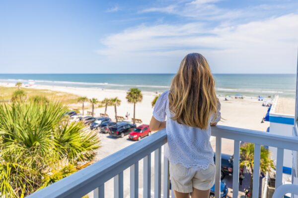 A young woman with long blonde hair stands on a balcony overlooking a sunny beach, palm trees, parked cars, and the ocean under a blue sky with scattered clouds.