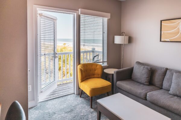 A cozy living room with a gray sofa, yellow armchair, white coffee table, and floor lamp. An open door and windows reveal a balcony with a view of the ocean and palm trees outside.