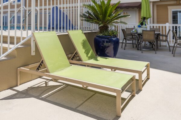 Two green lounge chairs sit side by side on a poolside patio near stairs, with a large potted plant and a table with chairs and an umbrella in the background.