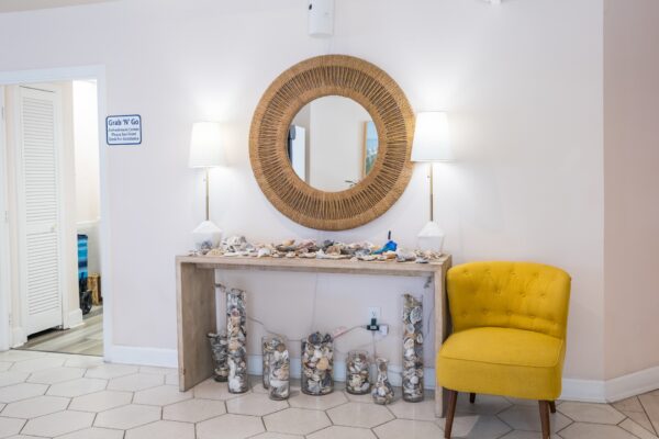 A console table decorated with seashells and glass vases sits against a white wall, beneath a round wicker mirror and two white lamps; a yellow armchair is on the right side of the table.