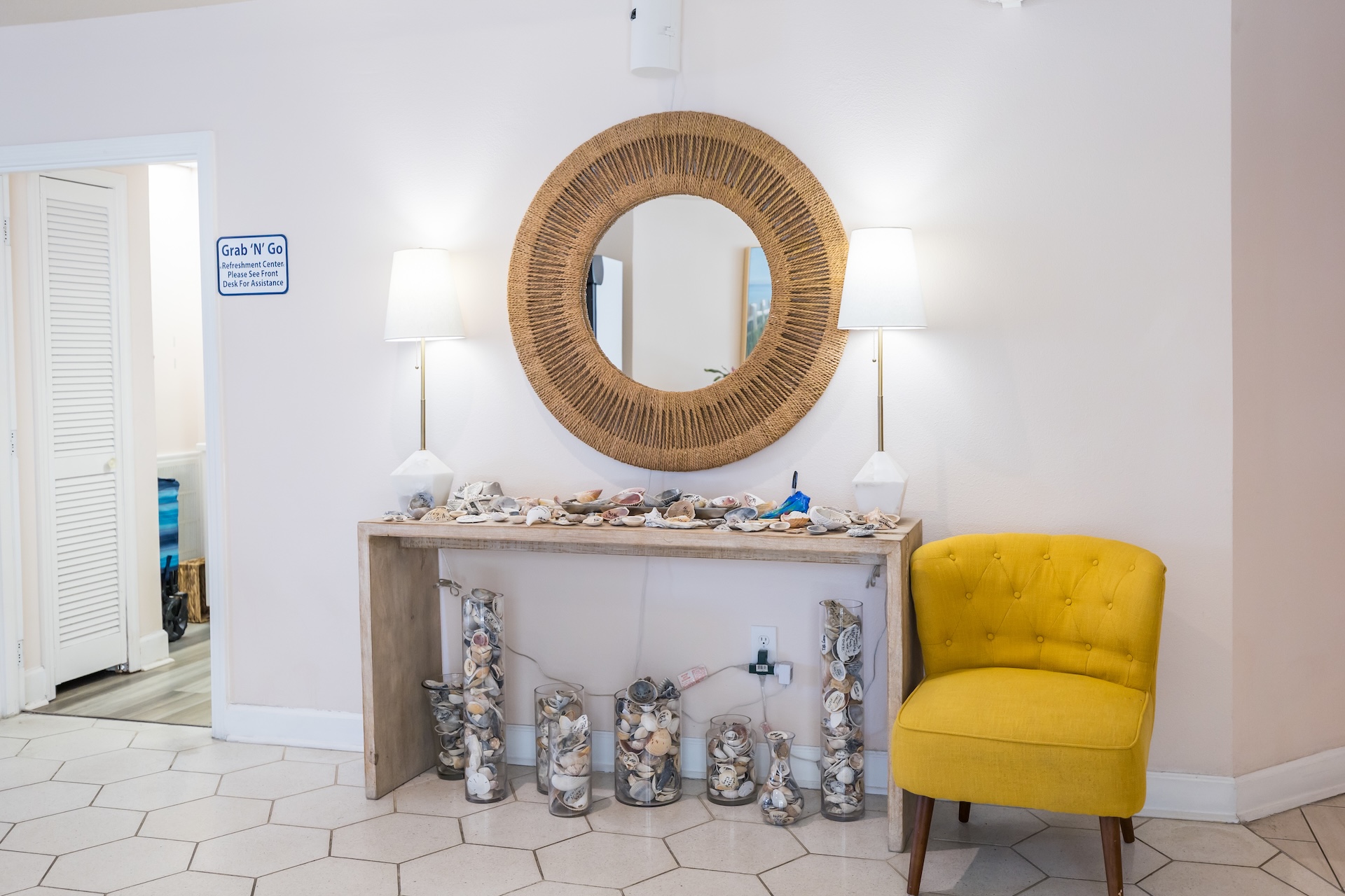 A console table decorated with seashells and glass vases sits against a white wall, beneath a round wicker mirror and two white lamps; a yellow armchair is on the right side of the table.