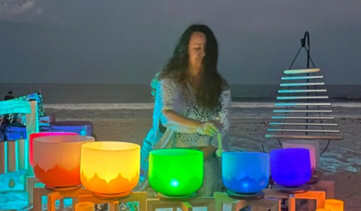 A person with long hair sits on a beach at dusk, playing colorful crystal singing bowls arranged in a rainbow order, with candles and a chime instrument nearby, as waves are visible in the background.