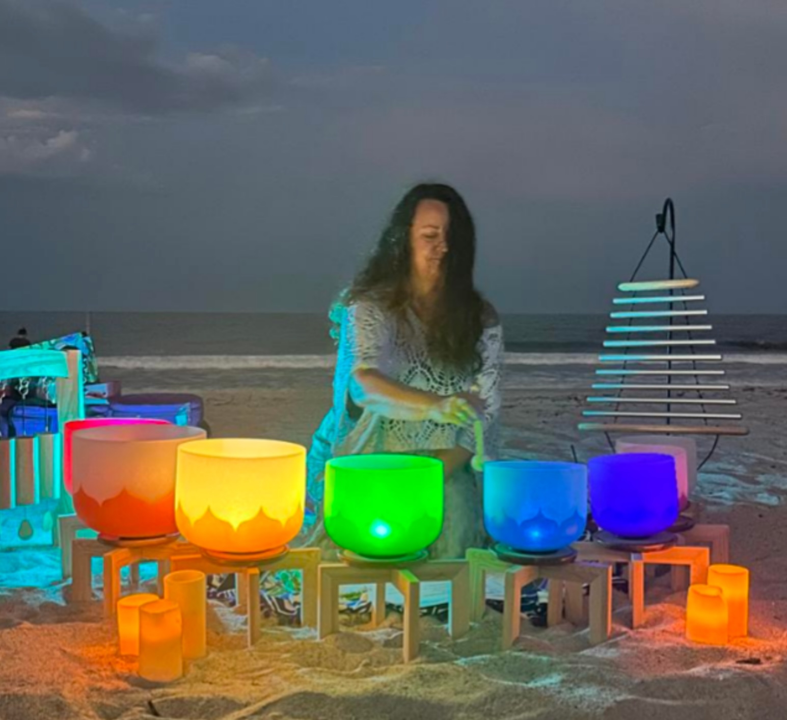 A person with long hair sits on a beach at dusk, playing colorful crystal singing bowls arranged in a rainbow order, with candles and a chime instrument nearby, as waves are visible in the background.