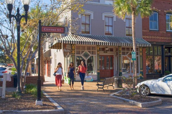 Three women walk along a sunny street with shopping bags near a sign reading Centre St. The area features palm trees, vintage-style buildings, and parked cars, creating a relaxed, small-town atmosphere.